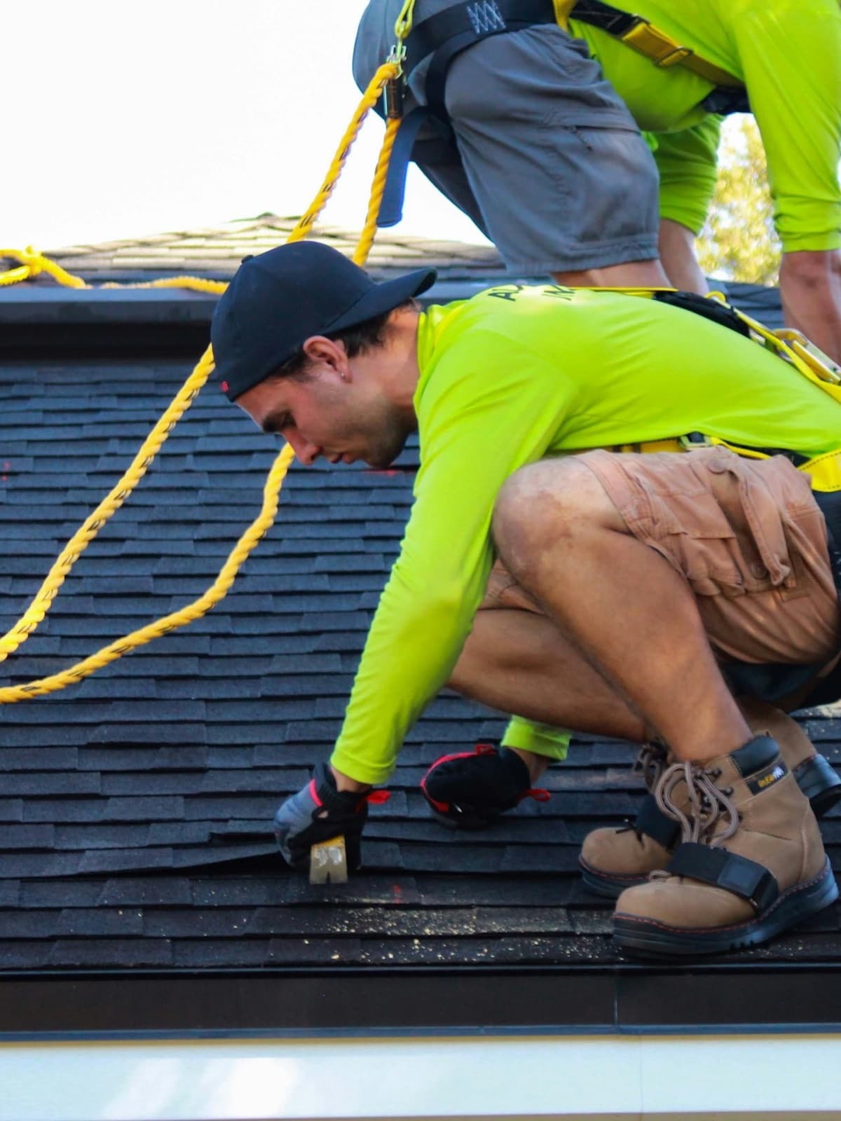 A roofer in a safety harness, cap and work gloves fixing shingles on a house roof